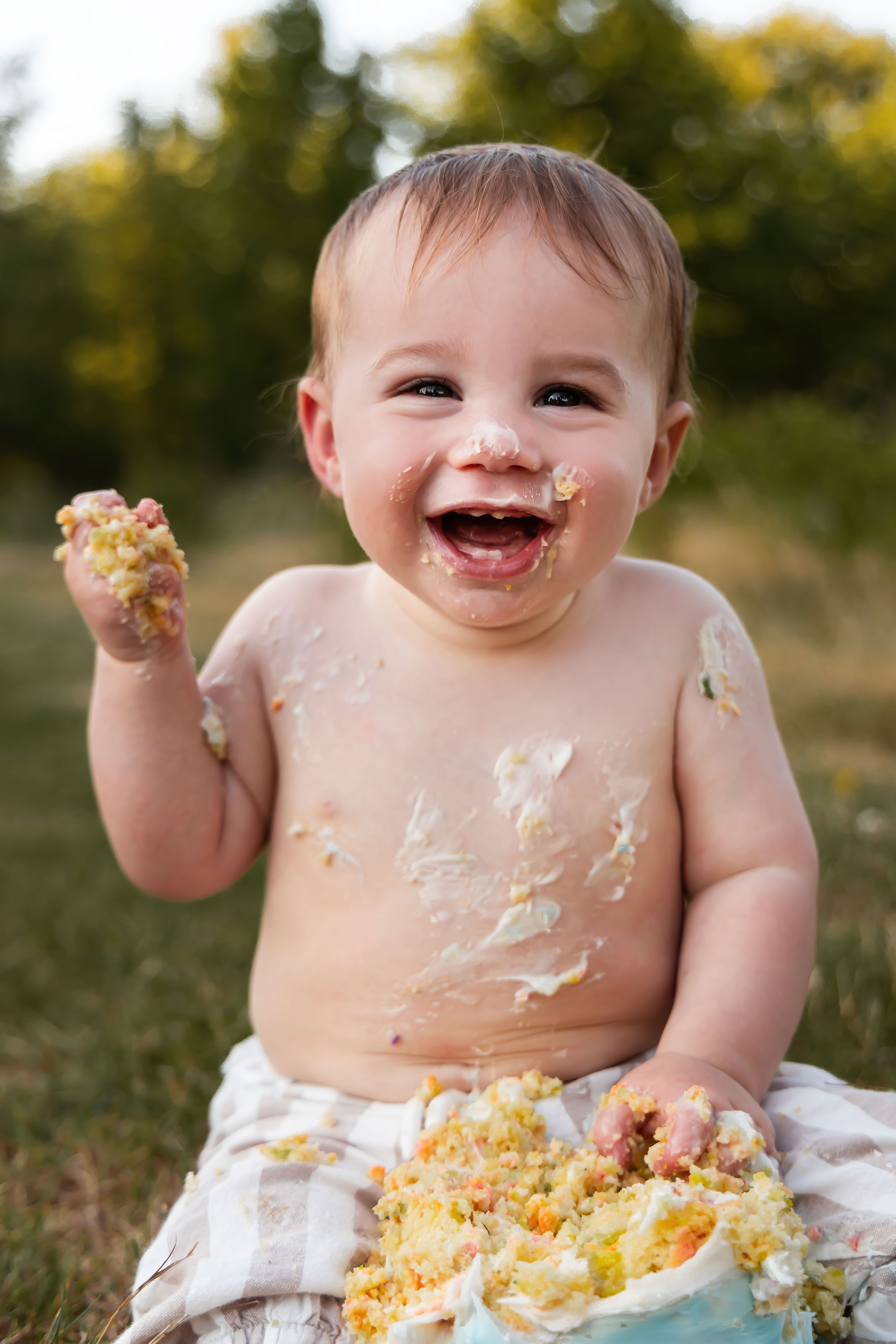 Natural light baby photography showcasing a happy first birthday cake smash in a beautiful outdoor setting with green trees.