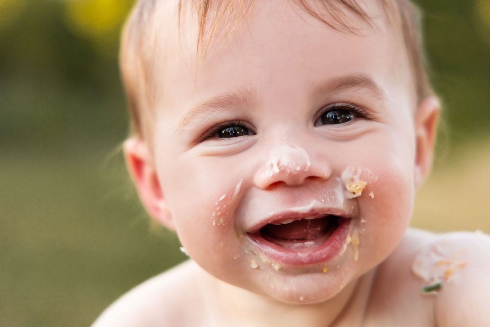 Fun outdoor first birthday portraits of a baby boy reaching toward the camera with cake and frosting on his arms.
