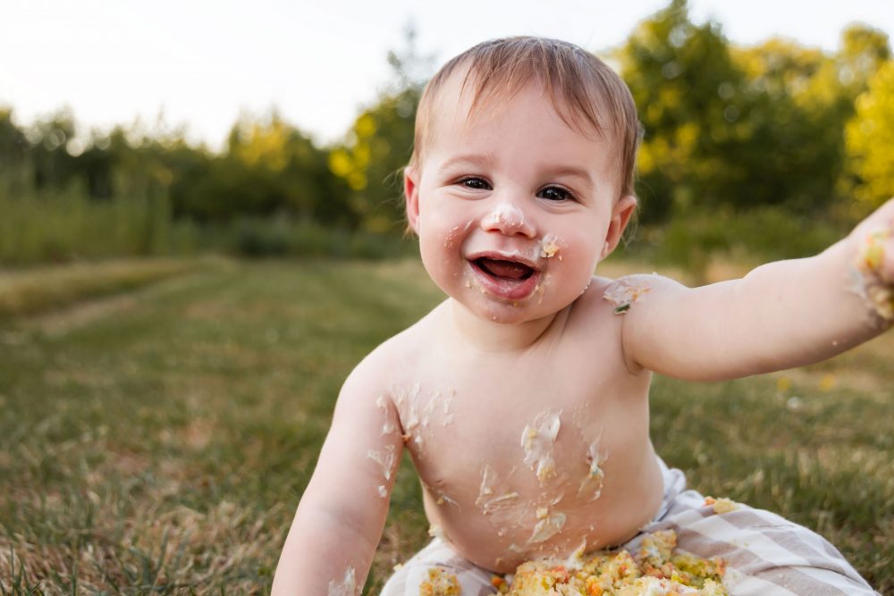 Fun outdoor first birthday portraits of a baby boy reaching toward the camera with cake and frosting on his arms.