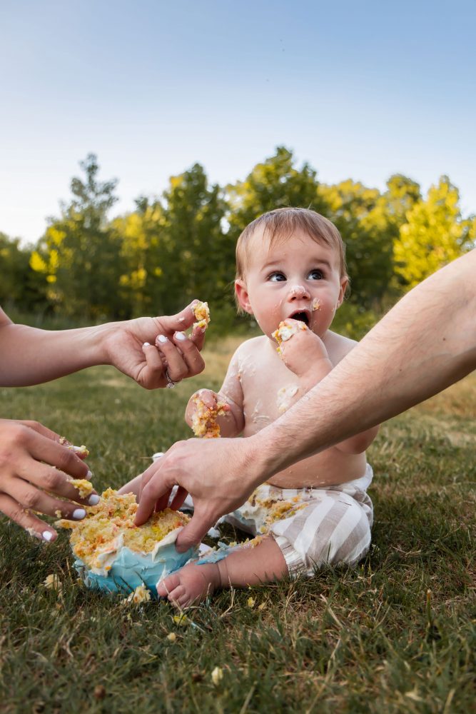 Natural light baby photography showcasing a happy first birthday cake smash in a beautiful outdoor setting with green trees.