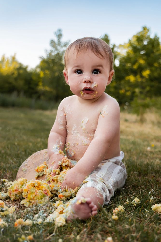 Happy one-year-old boy sitting in the grass with cake frosting on his face and tummy, holding a handful of colorful birthday cake.