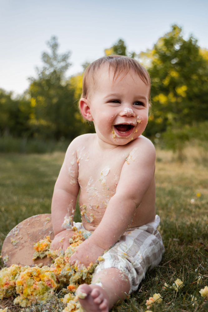Happy one-year-old boy sitting in the grass with cake frosting on his face and tummy, holding a handful of colorful birthday cake.